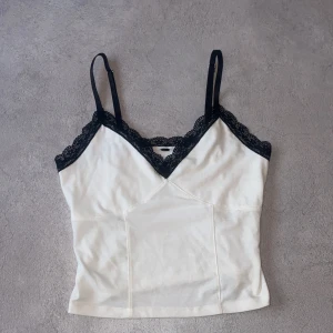 Vit linnetopp med spetsdetaljer - Säljer en stilren vit linnetopp med svarta spetsdetaljer längs urringningen. Toppen har justerbara axelband och en v-ringad design som ger en elegant touch. Perfekt för att lägga till en feminin känsla till din outfit.