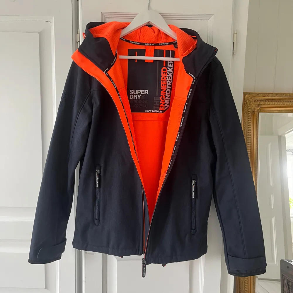 Jag säljer denna snygga jacka från Superdry. Jackan är av modellen hooded windtrekker och är en softshelljacka som är vind/vattenavvisande. Jackans utsida har en blågrå färg med orangea detaljer. Insidan är täckt med ett bekvämt innerfoder, som har en fin orange färg. Aldrig blivit använd och är därför i mycket fint skick utan defekter. Längst ner har jackan justerbara snören och även vid luvan. Pris kan diskuteras. Tveka inte att fråga om det är någonting du undrar över eller vid fler bilder!☺️. Takit.