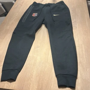Svarta mjukisbyxor från Nike med FC Barcelona-logga - Snygga svarta mjukisbyxor från Nike med FC Barcelona-logga på ena benet och guldfärgad Nike-swoosh på andra. Byxorna har snörning i midjan och en dragkedjeficka på sidan. Perfekta för fotbollsfans!