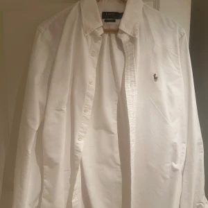 Vit skjorta från Ralph Lauren - Klassisk vit skjorta från Ralph Lauren med broderad logga på bröstet. Skjortan har en normal passform och är långärmad med knappar framtill. Perfekt för en stilren look.
