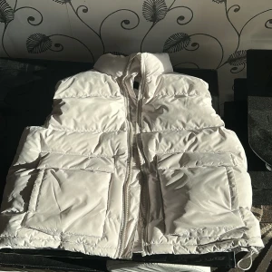 Vit dunväst från H&M - Säljer en stilren vit dunväst från H&M. Västen har en dragkedja framtill och två stora fickor. Perfekt för kyliga dagar med sin vattenavvisande och vindtäta funktion.