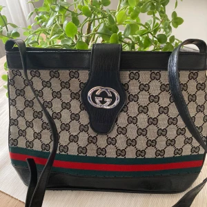 Mönstrad axelväska från Gucci - Snygg axelväska från Gucci med det klassiska GG-mönstret. Väskan har svarta skinnremmar och detaljer, samt en ikonisk logotyp framtill. Den har en dragkedja upptill och ett rymligt inre med extra fack. Perfekt för att ge din outfit en lyxig touch.