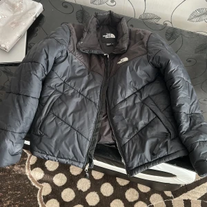 Svart pufferjacka från The North Face - Säljer en snygg svart pufferjacka från The North Face. Jackan har en dragkedja framtill och är perfekt för kyligare dagar. Den har långa ärmar och en klassisk design med logotypen på bröstet. Perfekt för dig som vill hålla stilen i vinter! 🖤 
