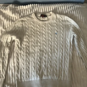 Vit kabelstickad tröja från Morris - Säljer en stilren vit kabelstickad tröja från morris .Tröjan har en klassisk rund halsringning och ett diskret blått broderat emblem på bröstet. Perfekt för en elegant och avslappnad look. Kan vara på gränsen till urtvättad men inte några fläckar eller andra synliga grejer.