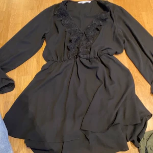 Svart klänning med spetsdetaljer - Snygg svart klänning med långa ärmar och v-ringning prydd med spetsdetaljer. Perfekt för en stilren look.