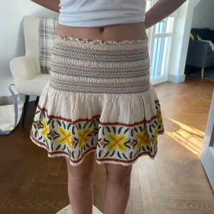 Blommig kjol med volang - Charmig kortkjol med volang och färgglada blommor. Elastisk midja med smockdetaljer och broderade blommor i gult och orange. Perfekt för sommarens äventyr!