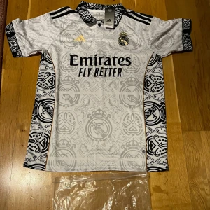 Vit fotbollströja från Adidas - Säljer en helt ny och oandvänd vit fotbollströja från Adidas med svart och guld detaljer. Tröjan har ett unikt mönster och korta ärmar. Perfekt för fotbollsfans som vill ha något speciellt.