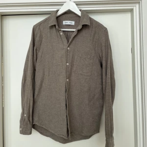 Flanellskjorta från Samsøe Samsøe - Snygg beige flanellskjorta  från Samsøe Samsøe med klassisk krage och knappar framtill. Skjortan har en bröstficka och är tillverkad i ett mjukt material som ger en avslappnad look. Perfekt för en stilren vardagsoutfit. Skick enligt bilder.