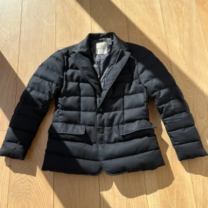 Svart quiltad kavaj från Moncler - Snygg svart quiltad kavaj från Moncler med knappar framtill. Jackan har en stilren design och är varm och skön, jackan har en liten lagning vid ena ärmen som inte syns. Nypris 8k