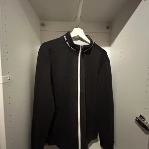Svart hoodie från Hugo Boss - Snygg svart jacka från Boss med vit dragkedja och textdetaljer på kragen. Perfekt för en stilren look. Jackan har långa ärmar och en normal passform.