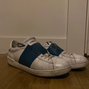 Valentino Garavani sneakers i vitt och blått - Snygga Valentino Garavani sneakers i vitt med breda blå detaljer. Skorna har en klassisk design med snörning och en bekväm passform. Perfekta för en stilren look.