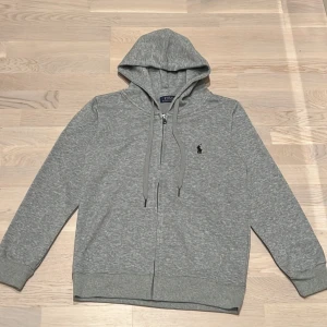 Grå hoodie från Ralph Lauren - Säljer en stilren grå hoodie från Ralph Lauren med dragkedja och justerbar huva. Den är dock liten i storleken.