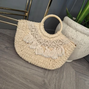 Boho handväska  - Charmig beige handväska i flätat material med rundade handtag. Väskan har dekorativa fransar och ett vackert mönster på framsidan. Perfekt för en bohemisk stil.