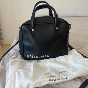 Svart handväska från Balenciaga - Snygg svart handväska från Balenciaga med stilren design. Väskan har en praktisk dragkedja och ett rymligt rött innerfoder med flera fack. Perfekt för att bära med sig det viktigaste med stil. väskan är i nyskick.