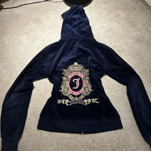 Mörkblå velourjacka från Juicy Couture - Säljer en snygg mörkblå velourjacka från Juicy Couture med dragkedja och huva. Jackan har ett stort, detaljerat broderi på ryggen med guld- och rosa detaljer samt en liten broderad logga på framsidan. Perfekt för en avslappnad och stilren look.