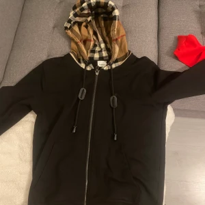 Burberry hoodie - Säljer en svart hoodie med dragkedja och rutigt foder i huvan. Huvan har snörning och jackan har långa ärmar. Perfekt för en avslappnad stil.