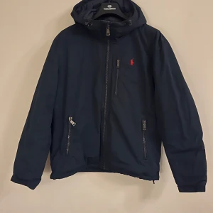 Mörkblå jacka från Ralph Lauren - Säljer en snygg mörkblå jacka från Ralph Lauren med huva och dragkedja. Jackan har två praktiska fickor med dragkedjor och en liten röd logga på bröstet. Perfekt för kyligare dagar.
