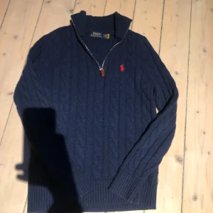 Mörkblå stickad tröja från Polo Ralph Lauren - Säljer en snygg mörkblå stickad tröja från Polo Ralph Lauren med dragkedja och röd logga på bröstet. Tröjan har en ribbad krage och långa ärmar, perfekt för kyliga dagar.lite nopprig och sitter som xs