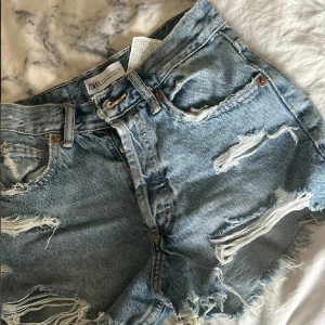 Blå jeansshorts från Zara - Säljer ett par blå jeansshorts från Zara med slitna detaljer och fransar. Perfekta för en avslappnad sommarlook. De har en klassisk femficksdesign och knappgylf.
