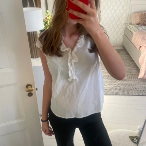 Vit blus från Holly & White - Säljer en söt vit blus från Holly & White med volangdetaljer. Blusen är ärmlös och har en luftig passform, perfekt för varmare dagar. Den är gjord i ett mjukt och bekvämt material.