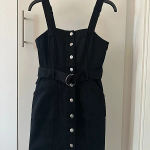 Svart klänning med knappar och bälte - Snygg svart klänning med knappar framtill och ett matchande bälte i midjan. Klänningen har breda axelband och en stilren design som passar perfekt för olika tillfällen. Material likna jeanstyg