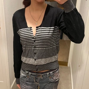 Svart och grå randig kofta från Lindex - Säljer en snygg svart och grå randig kofta .Koftan har en design med knappar framtill och långa ärmar. Perfekt för en stilren look!