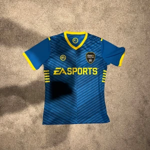 FIFA 17 Ultimate Team fotbollströja - Säljer en blå fotbollströja från EA Sports med gula detaljer och korta ärmar. Tröjan har ett diagonalt randigt mönster och nummer 17 på ryggen. Perfekt för fotbollsfans som vill sticka ut på planen!