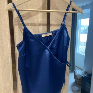 Blå omlott-topp från NA-KD - Snygg blå topp från NA-KD med smala axelband och knytning i sidan. Satinliknande material.