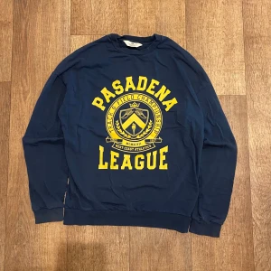 Mörkblå Sweatshirt Från H&M Storlek 170 - M - Säljer en mörkblå sweatshirt från H&M med tryck i gult som säger 'Pasadena League'. Tröjan har långa ärmar och en rund halsringning. Perfekt för en avslappnad stil. Storlek 170 Passar Medium. Snabb Leverans 