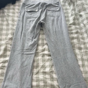 Grå juicy couture - Säljer ett par grå mjukisbyxor med bred resår i midjan och snörning framtill. Byxorna har två stora fickor fram och två bakfickor med knappar. Perfekta för en avslappnad stil.