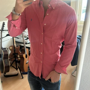 Rosa skjorta från Ralph Lauren - Snygg rosa skjorta från Ralph Lauren med klassisk krage och knappar framtill. Den har en broderad logga i blått på bröstet. Perfekt för en stilren look.