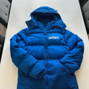 Blå pufferjacka från Hoodrich - Säljer en blå pufferjacka från Hoodrich med broderad logga på bröstet och ryggen. Jackan har en dragkedja framtill och långa ärmar. Perfekt för kyliga dagar med sin varma och vadderade design.