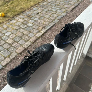Svarta fotbollsskor från Nike - Snygga svarta fotbollsskor från Nike med vita detaljer och blå inslag. Skorna har snörning och en slitstark sula med dobbar för bra grepp på planen. Perfekta för fotbollsträning och matcher.