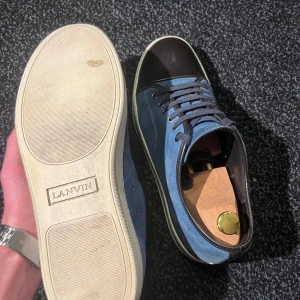 Blå sneakers från Lanvin - Snygga blå sneakers i mocka från Lanvin med mörkblå detaljer och snörning. Skorna har en stilren design med en kontrasterande svart tå och en ljus sula. Perfekta för en trendig look.     Skorna är i bra skick och i storlek EU 43/ UK 9. Dustbag ingår ✅.                                               Tveka inte att höra av er vid frågor!