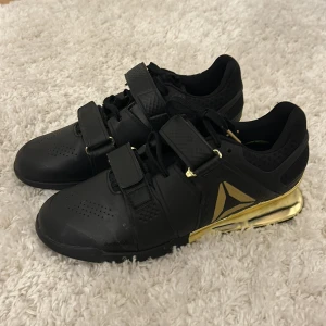 Svarta tyngdlyftare skor med gulddetaljer från Reebok - Snygga svarta träningsskor från Reebok med gulddetaljer. (Använd fåtal gånger)