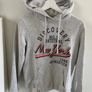 Grå hoodie från Twintip - Säljer en grå hoodie från Twintip med tryck 'Discovery New York' i svart och rosa. Tröjan har en justerbar huva med dragsko och långa ärmar. Perfekt för en avslappnad stil. Precis som ny! Storlek S