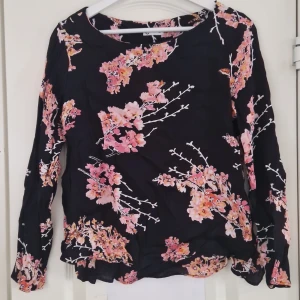 Svart blommig blus - Säljer en svart blus med vackert blommönster i rosa och orange nyanser. Blusen har långa ärmar och en rund halsringning. Perfekt för att ge en färgklick till din garderob!