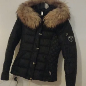 Svart dunjacka från Rockandblue - Säljer en snygg svart dunjacka från Rockandblue med en stor, fluffig pälsdetalj runt huvan. Jackan har en quiltad design och stängs med både dragkedja och knappar. Perfekt för kalla dagar när du vill hålla stilen. 🖤