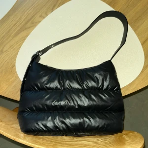 Vadderad axelväska - Svart axelväska i nylon med quiltad design. Väskan har en justerbar axelrem med silverfärgade detaljer. Stängs med dragkedja. Säljes i nyskick! ✨