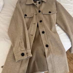 Snygg beige kappa med svarta knappar och knytband i midjan. Den har långa ärmar och två bröstfickor med lock. Perfekt för höst och vår! 