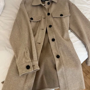 Beige kappa med knappar - Snygg beige kappa med svarta knappar och knytband i midjan. Den har långa ärmar och två bröstfickor med lock. Perfekt för höst och vår! 