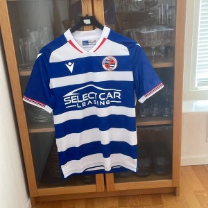 Reading FC hemmatröja 24/25 Macron S - Reading FC:s officiella hemmatröja 2024/25 från Macron. Tröjan är kortärmad och har breda blå och vita horisontella ränder, klubbmärke på bröstet och sponsortryck framtill. Tillverkad i tekniskt funktionsmaterial som andas. Snygga röda detaljer vid krage och ärmslut.