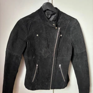 Svart läderjacka - Snygg svart läderjacka med asymmetrisk dragkedja och coola detaljer. Jackan har två fickor med dragkedjor och en hög krage med tryckknapp. 