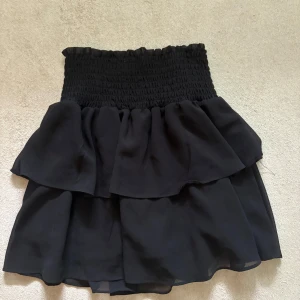 Svart volangkjol från Chelsea - Supersöt svart kortkjol från Chelsea med bred smock i midjan och dubbla volanger. Kjolen är luftig och har en flowy känsla, perfekt för dig som gillar en feminin och trendig stil. Materialet känns lätt och passar grymt till sneakers eller boots.