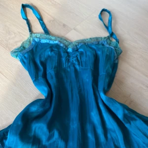 Turkos kortklänning med spetsdetaljer - Snygg turkos kortklänning i glansigt material med smala axelband och vackra spetsdetaljer upptill och nertill. Klänningen har en lätt utställd form och broderad spetskant som ger en romantisk vibe. Perfekt för dig som vill sticka ut med färg och detaljer.