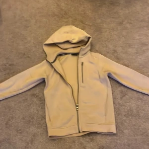 Beige Sail Racing fleecejacka med huva - Beige fleecejacka från Sail Racing med huva och hel dragkedja framtill. Jackan har en bröstficka med dragkedja och är långärmad. Perfekt för kyliga dagar och enkel att matcha med andra plagg.