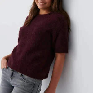 Mörk vinröd stickad tröja kort ärm - Mysig mörk vinröd stickad tröja med korta ärmar och rund hals. Tröjan har en lite loose passform och är perfekt att styla med jeans eller kjol. Materialet är fluffigt och ger en trendig look, perfekt för höst och vinter.