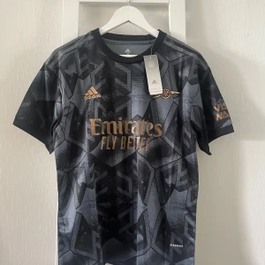 Arsenal svart bortatröja Adidas M - Snygg Arsenal bortatröja från Adidas i svart med geometriskt mönster och detaljer i guld. Kortärmad modell med rund hals och tryckta loggor som Emirates och Visit Rwanda. Tillverkad i lätt och ventilerande Aeroready-material.