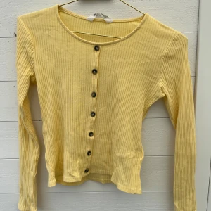 Gul ribbad topp med knappar - Snygg ljusgul ribbad topp från H&M med långa ärmar och knappar framtill. Toppen har en rund halsringning. Jättefin till våren o påsken. Passar mig som brukar ha XS