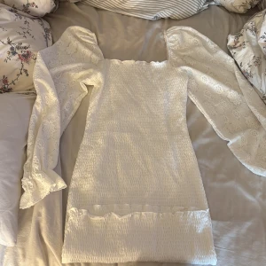 Vit kort klänning från Bubbleroom - Supersöt vit kort klänning från Bubbleroom med puffiga långärmade spetsärmar och smockad kropp. Ärmarna har blommigt spetsmönster och klänningen har volangdetalj vid nederkanten. Perfekt för sommarens alla tillfällen! 